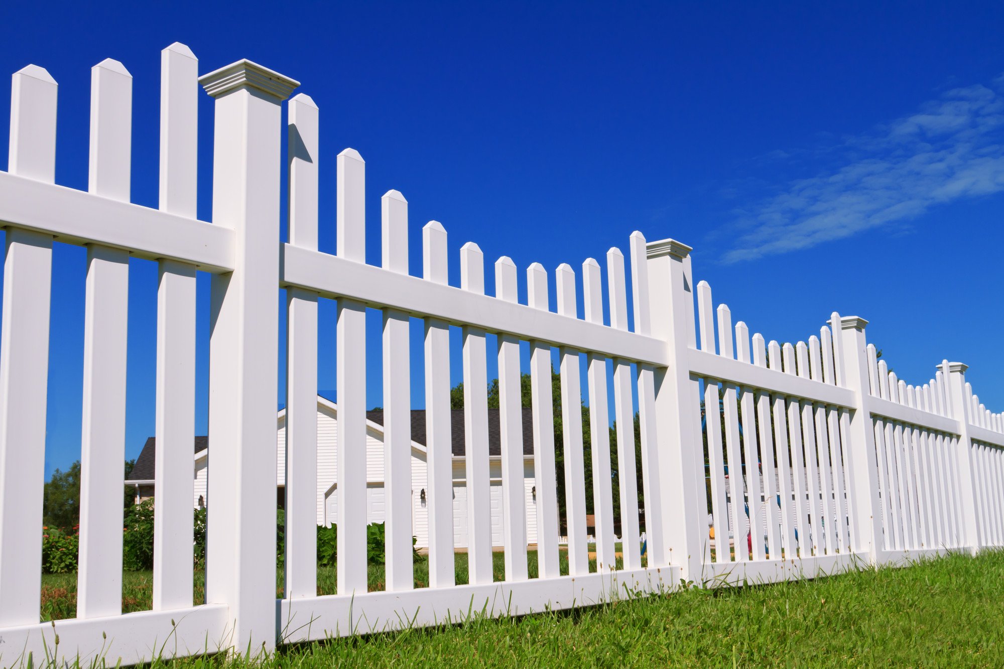 White vinyl picket fence with post caps running along a green lawn under a blue sky