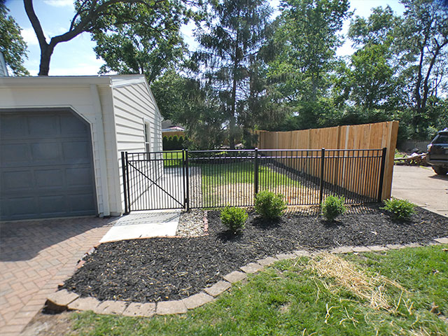 Black aluminum gate and fence closing the driveway side of the yard in Kirkwood, MO, with the existing wood privacy fence behind it