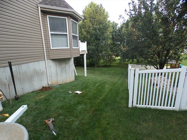 Side yard in Imperial before the fence, with the house wall on the left, an existing white fence on the right and tools and a fence post on the grass