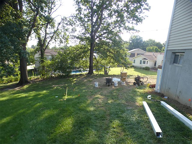 Backyard in Fenton before the fence, with marking stakes in the grass, a wheelbarrow, white vinyl rails on the ground and a large tree